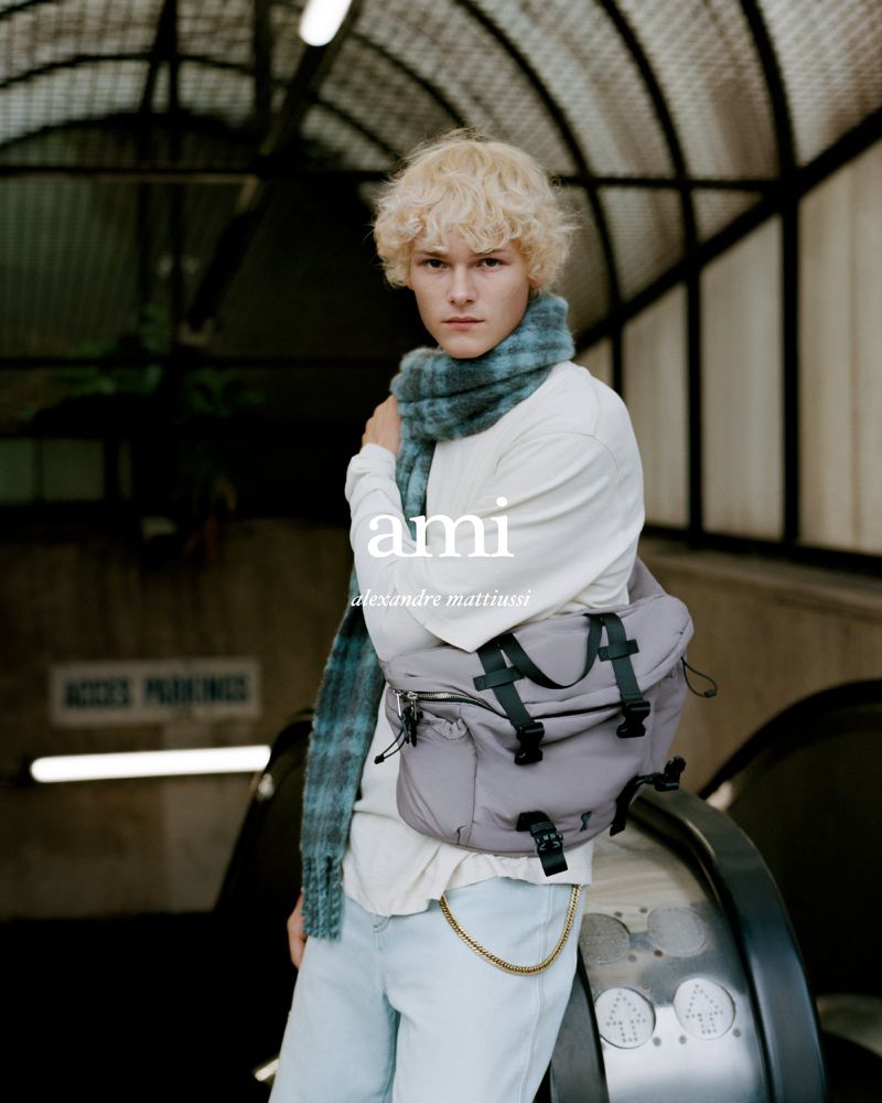 A blond male model poses in an underground tunnel wearing a scarf and carrying a grey messenger bag for a fashion advertisement.
