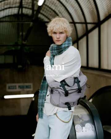A blond male model poses in an underground tunnel wearing a scarf and carrying a grey messenger bag for a fashion advertisement.