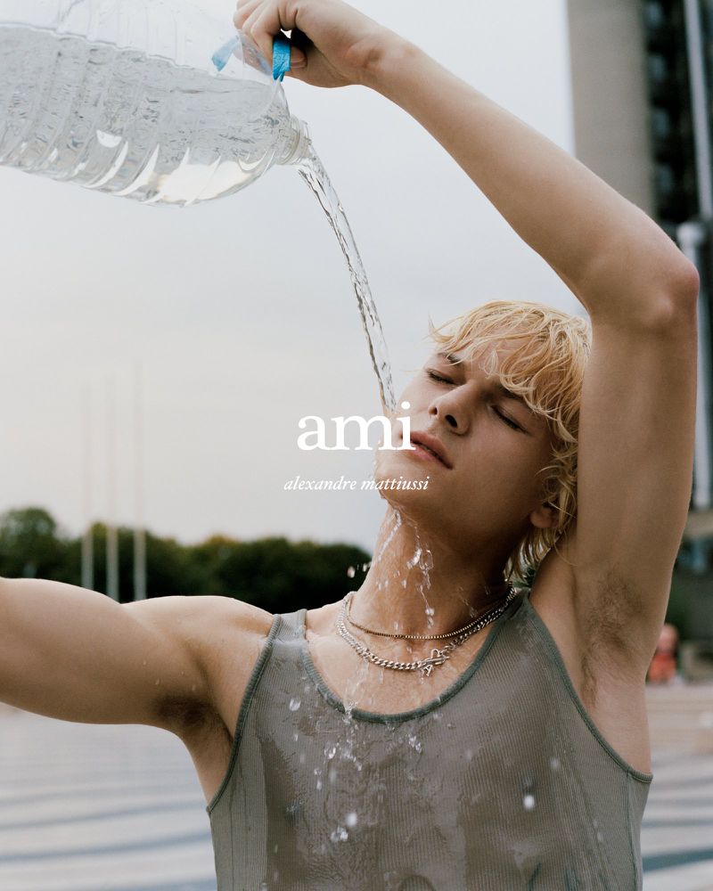 Blonde male model with short hair pours water on his face outdoors, wearing a semi-sheer tank top and layered necklaces, with AMI logo.