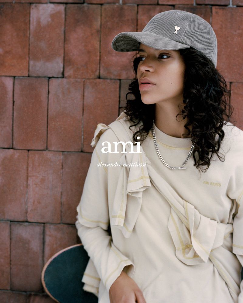 Woman wearing a gray corduroy baseball cap and beige sweatshirt stands next to a brick wall and skateboard in an outdoor setting.