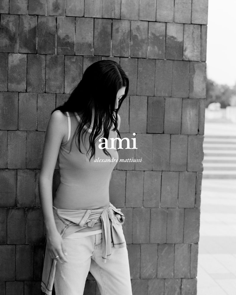 Black and white portrait of a woman in a tank top and jeans leaning against a brick wall with clothing brand logos, outdoors.