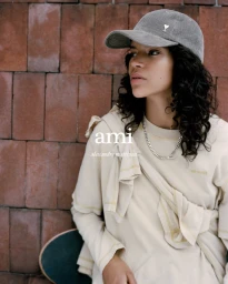 Model wearing a grey corduroy cap and cream sweatshirt leaning against a red brick wall with a skateboard
