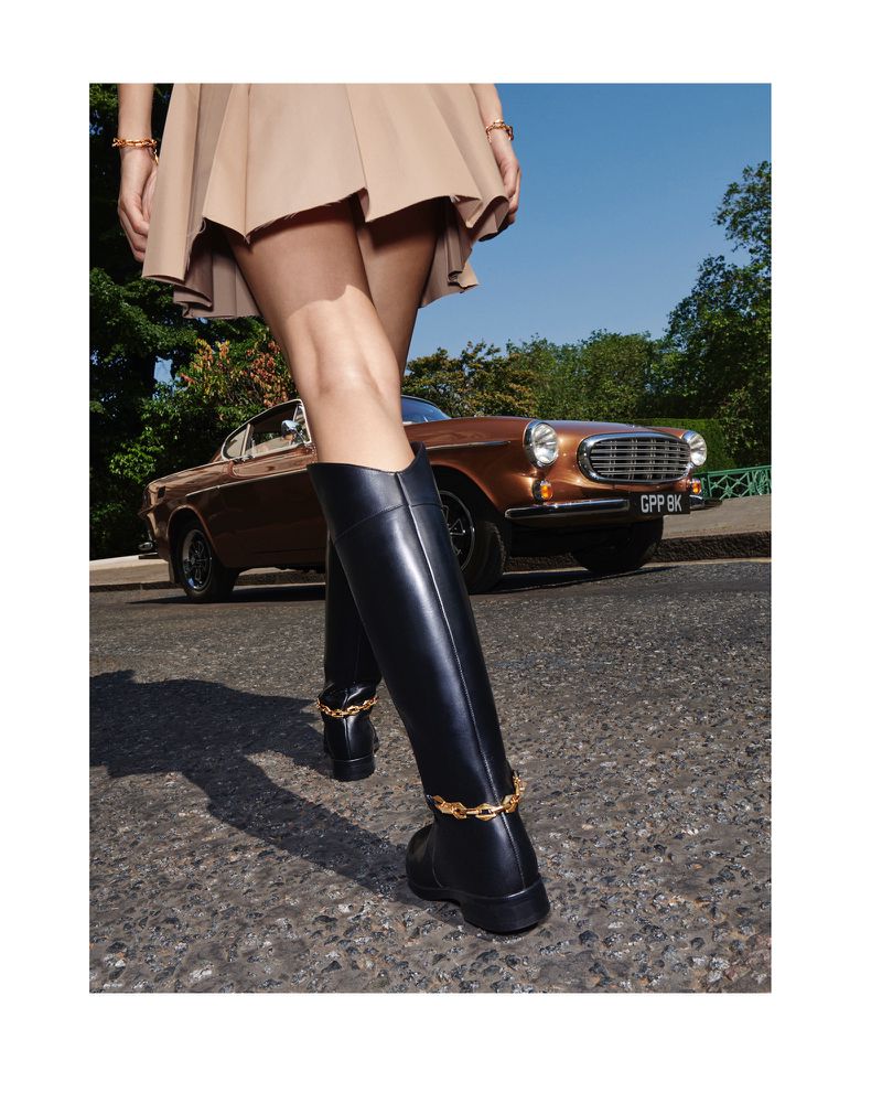 Model in tan skirt and black leather boots with gold ankle chain, posing on the street with vintage brown Volvo in the background.