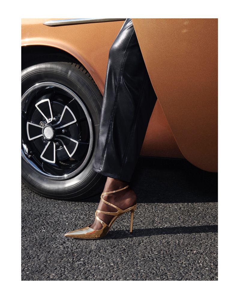 Dark-skinned person wearing black pants and gold high heels next to an orange car and black asphalt pavement. Fashion editorial.
