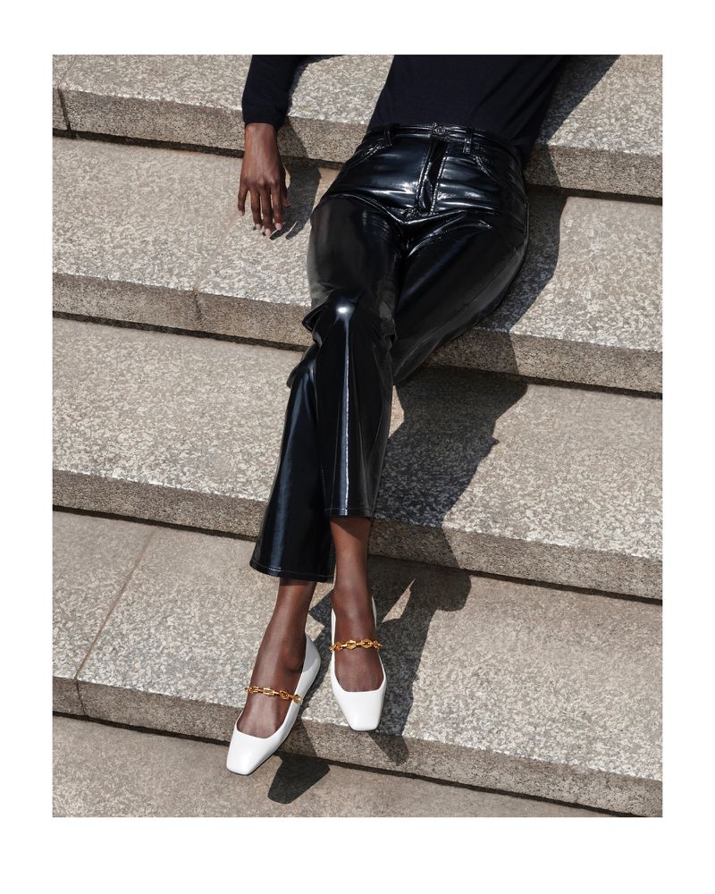 A woman poses on stone steps wearing black patent leather pants and white shoes with gold chain accents in a fashion-forward image.