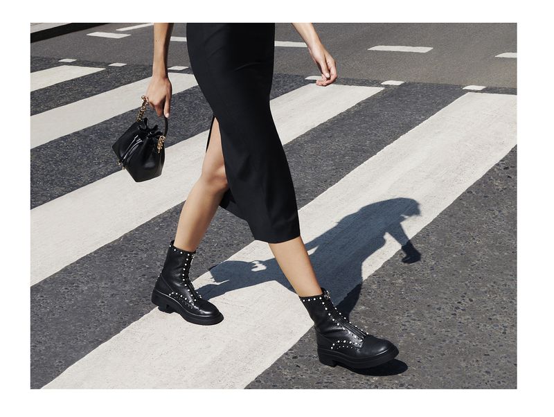 A woman walks across a crosswalk with a black dress, leather boots, and a purse, casting a shadow on the zebra-striped road.