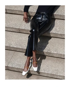 Model seated on stone steps wearing shiny black patent trousers and white shoes with gold chain straps.
