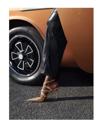 Close-up of metallic gold heeled shoe worn with black leather trousers beside a vintage car for Jimmy Choo.