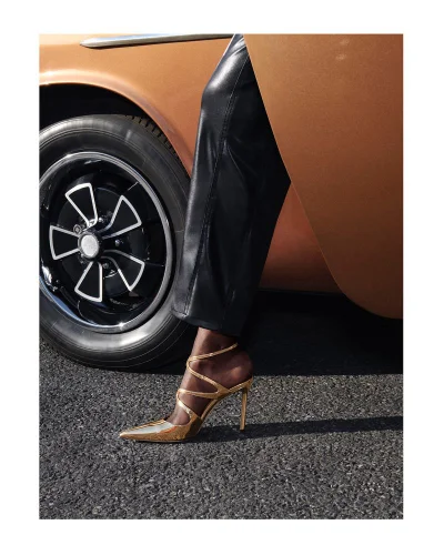 Close-up of metallic gold heeled shoe worn with black leather trousers beside a vintage car for Jimmy Choo.