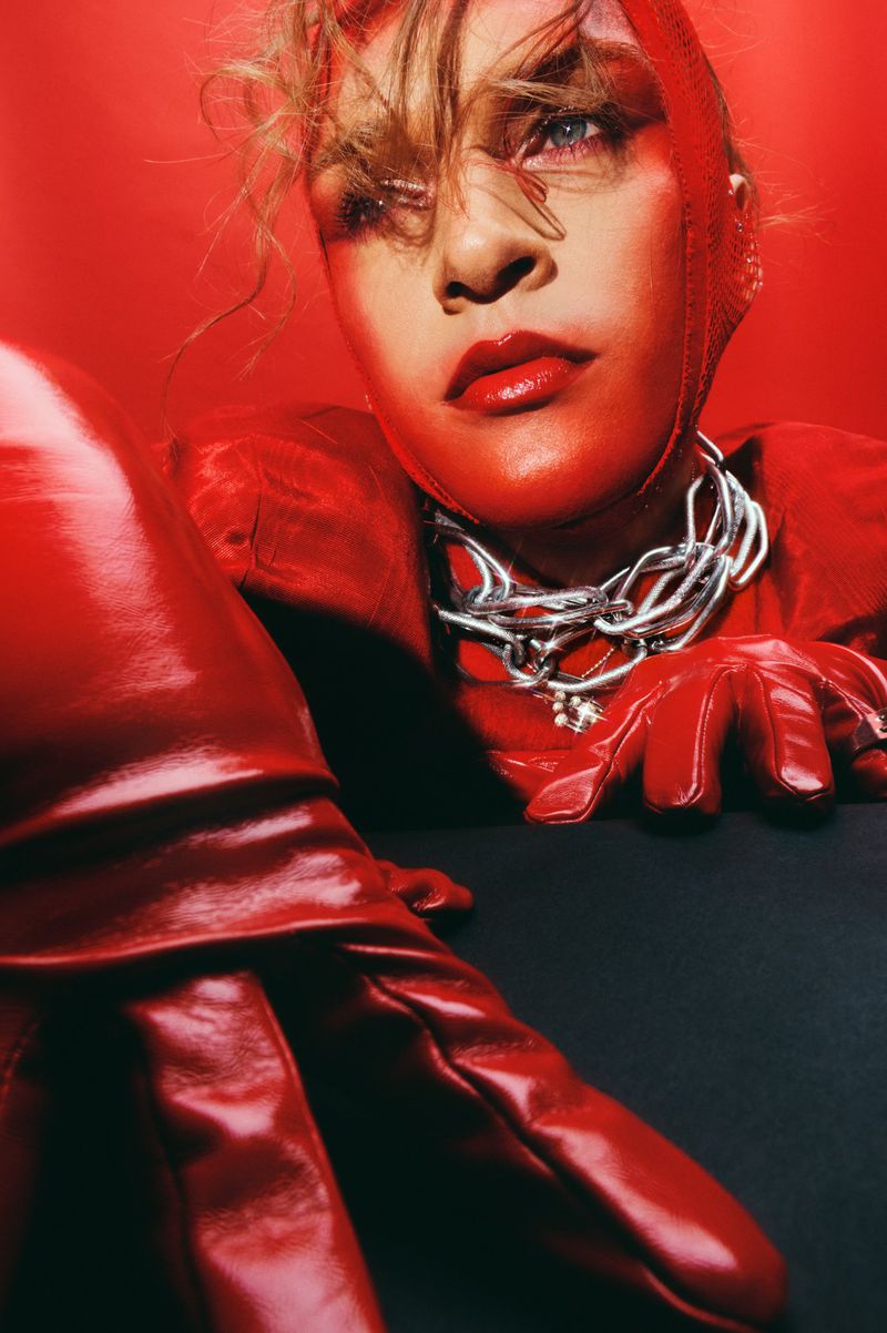 Woman in a red hood, gloves, and outfit with a silver chain, looking up against a matching red background in an eye-level shot.