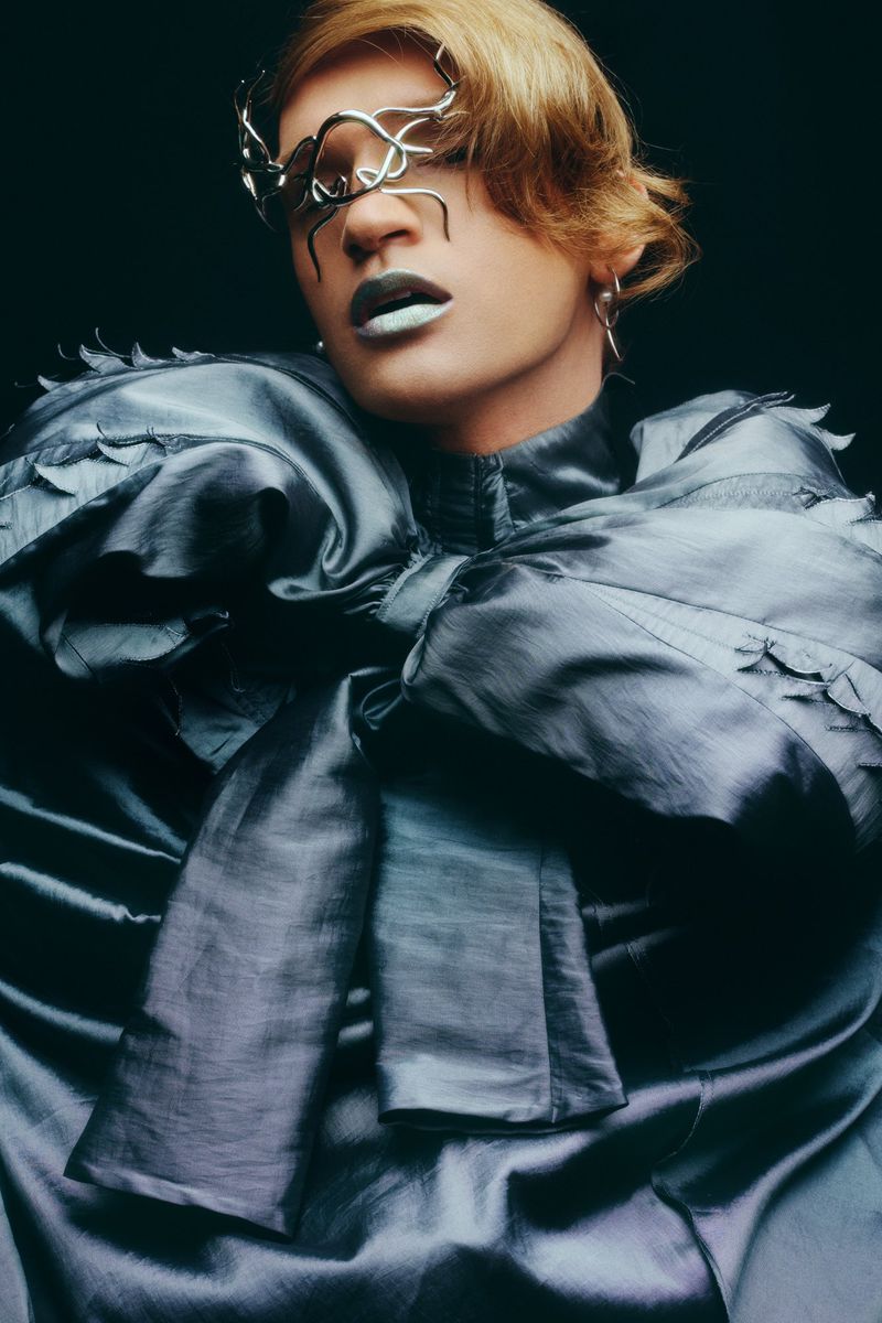 Close-up of a person with blond hair wearing a large grey satin garment and decorative eyewear against a dark backdrop in a studio.