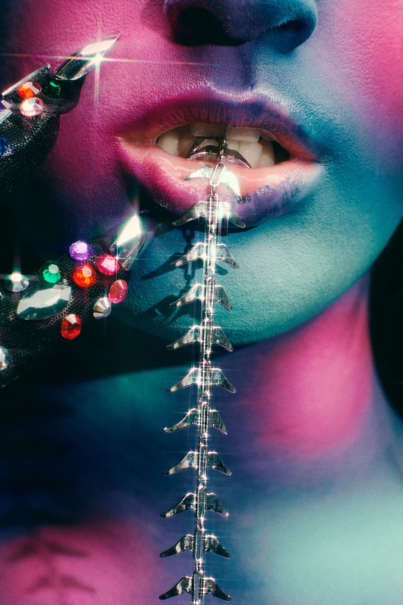 Close-up shot of a person with blue and pink makeup, adorned with jewel-encrusted jewelry and a spine-like ornament cascading from their mouth.