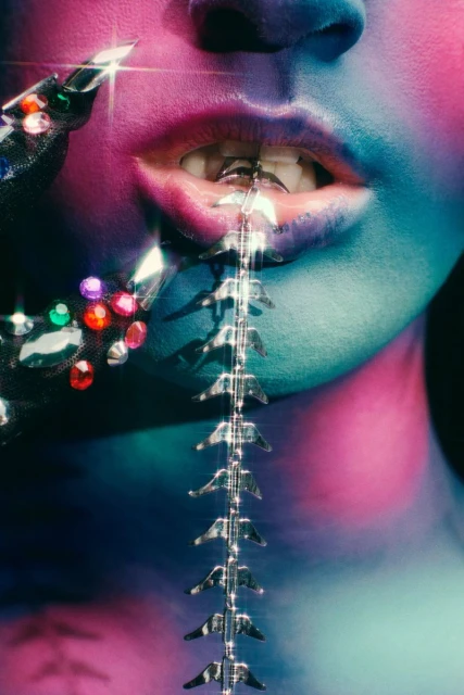 Close-up shot of a person with blue and pink makeup, adorned with jewel-encrusted jewelry and a spine-like ornament cascading from their mouth.