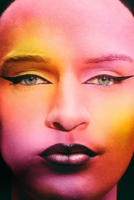 Close-up of a person with makeup including eyeliner and dark lipstick, featuring yellow, orange, pink, and red skin tones.