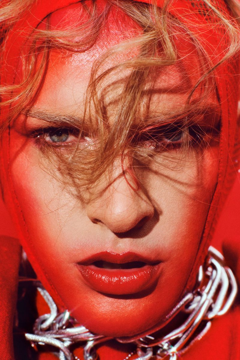 Close-up portrait of an adult woman with blonde hair, red makeup, wearing a silver chain necklace, and a red head covering.