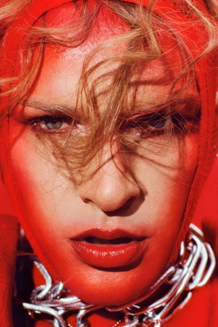 Close-up portrait of an adult woman with blonde hair, red makeup, wearing a silver chain necklace, and a red head covering.
