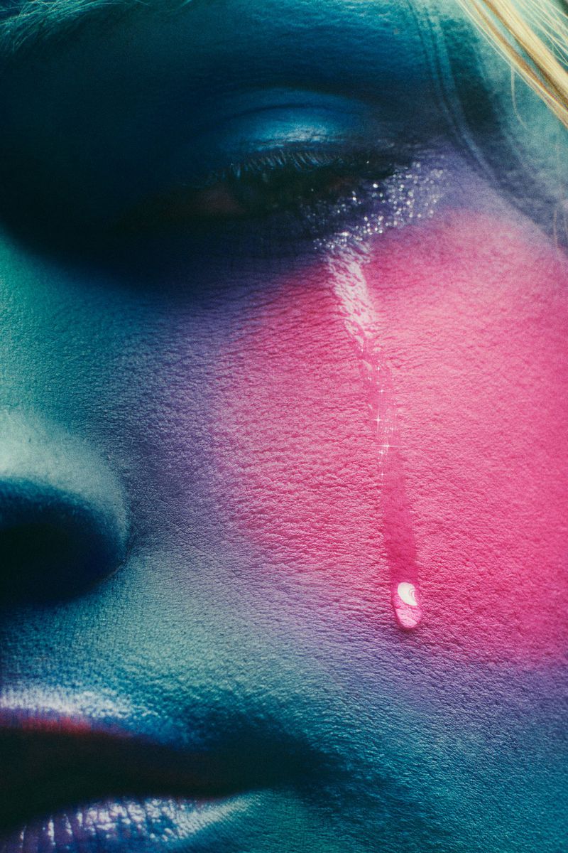 Close-up of a face with a visible tear running down the cheek. Vivid hues of pink, blue, and purple enhance the skin's texture.
