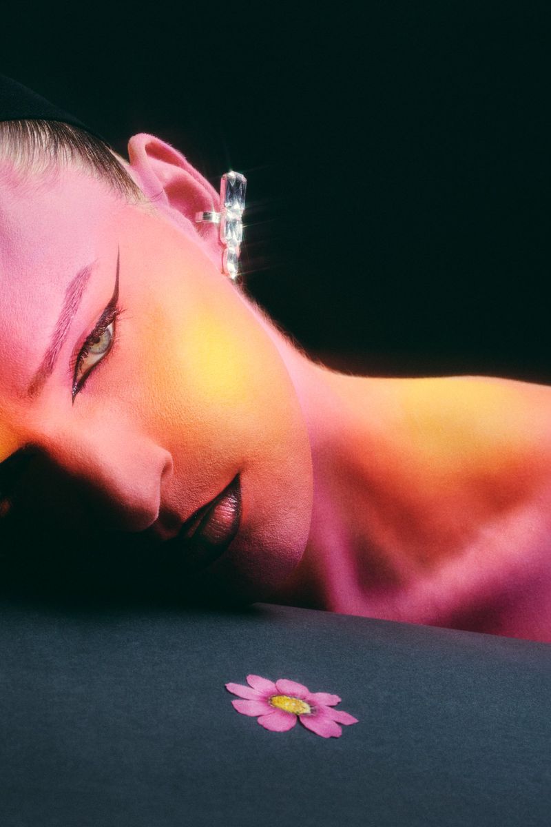 Close-up studio shot of a woman's face with pink and yellow lighting, dramatic makeup, crystal earring, and a single flower.