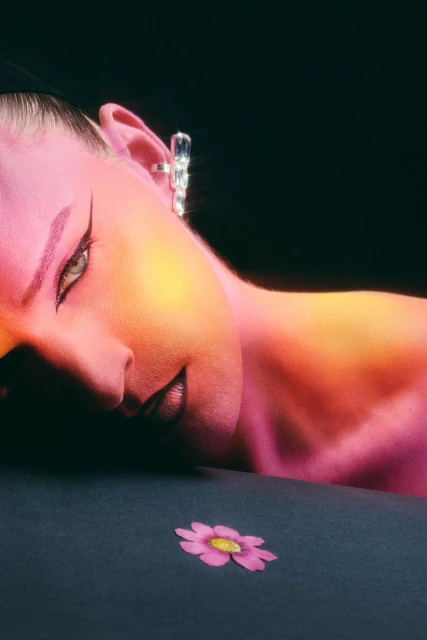 Close-up studio shot of a woman's face with pink and yellow lighting, dramatic makeup, crystal earring, and a single flower.
