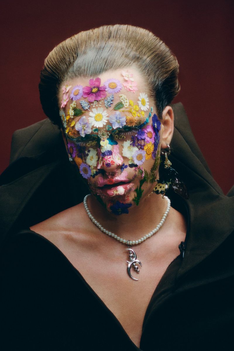 Person with face adorned with colorful flowers in a striking beauty shot with a pearl necklace and black top against a dark red background.