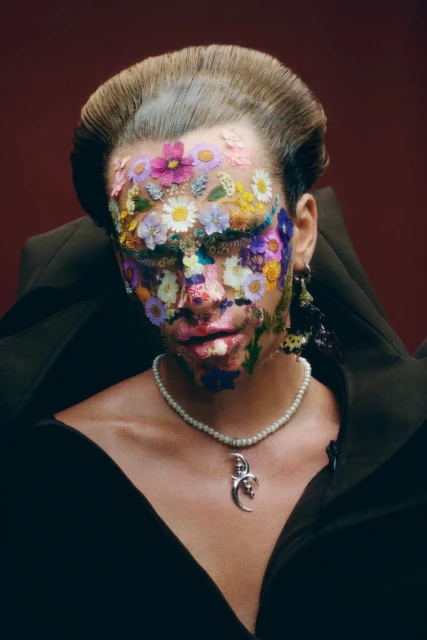 Person with face adorned with colorful flowers in a striking beauty shot with a pearl necklace and black top against a dark red background.