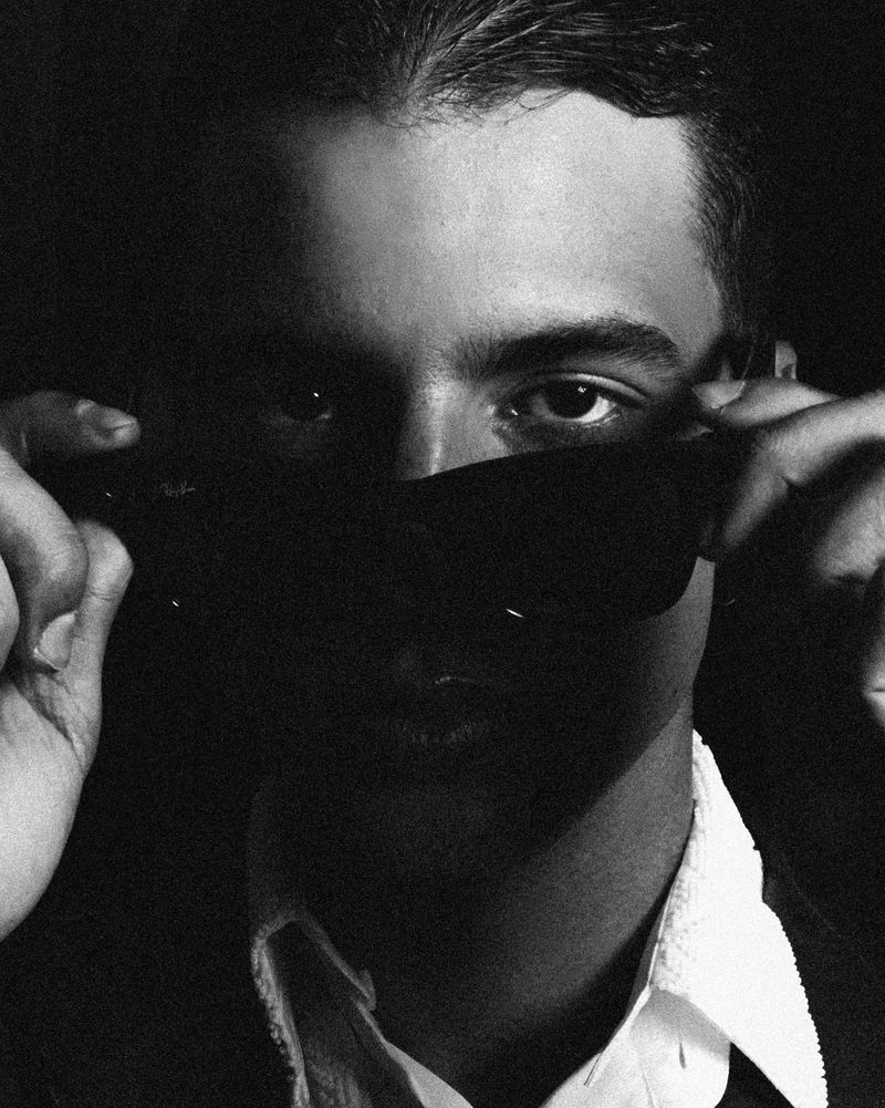 Monochrome portrait of a man partially obscured by sunglasses, set against a dark background, creating a dramatic and mysterious mood.