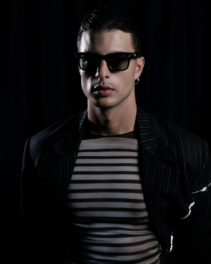 Portrait of a young adult male wearing sunglasses and a striped shirt under a blazer, set against a dark backdrop in a studio environment.