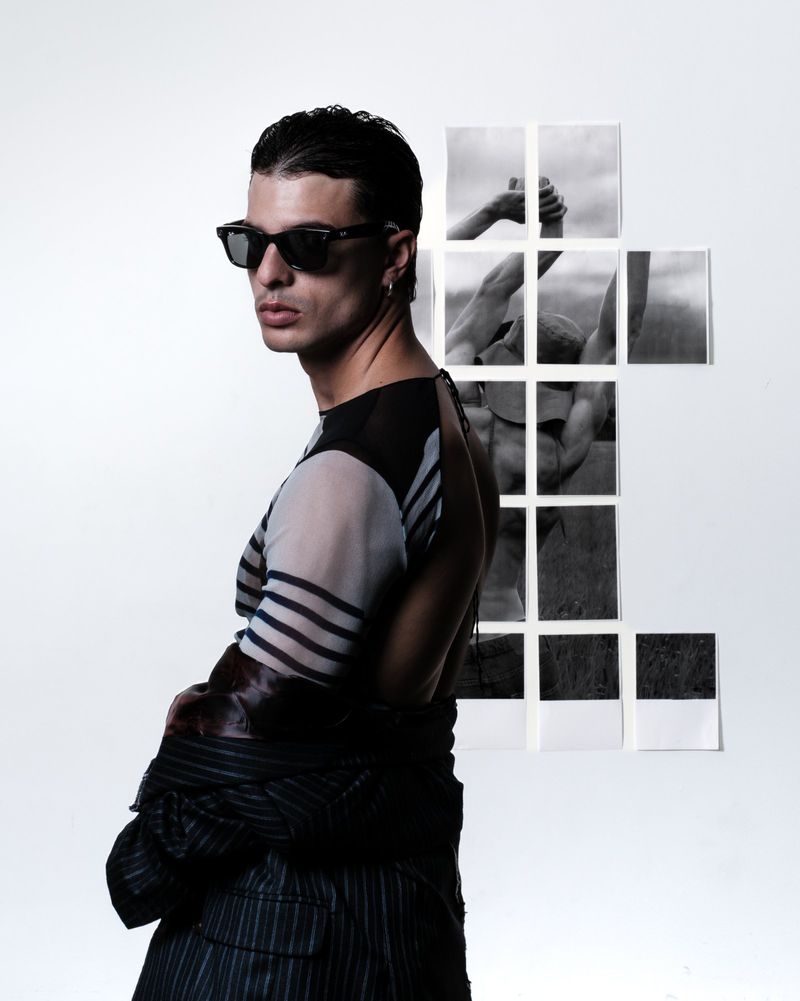 Stylish man in sunglasses poses in front of black-and-white photography. Dark hair, sheer top, jacket slung over shoulder. White background.