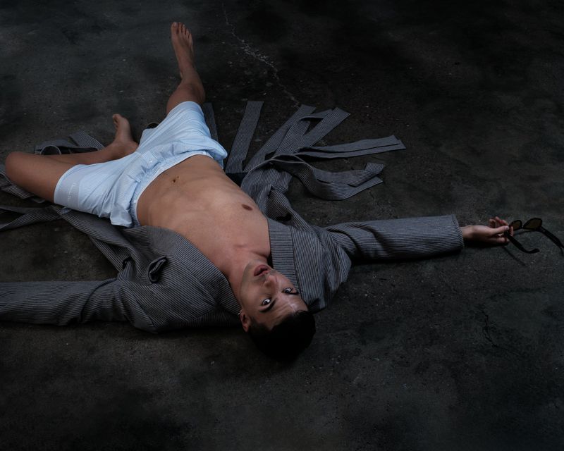 Young man laying on concrete floor wearing boxers and open robe holding glasses in an indoor fashion or beauty style photograph.
