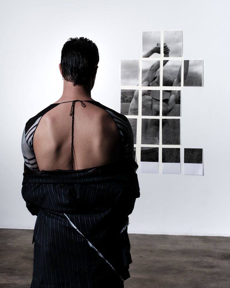 Back view of a slender adult male in an open pinstripe jacket looking at a collection of black-and-white photography in a white interior.