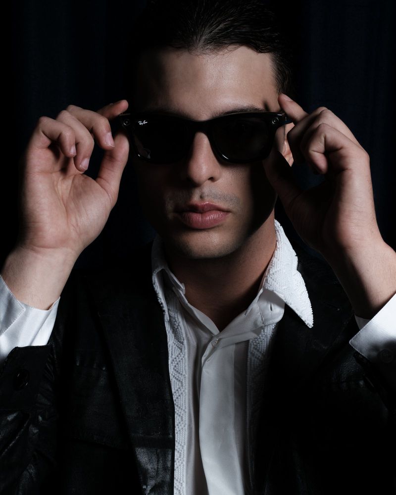 Portrait of a man wearing sunglasses and a black leather jacket indoors with a white button-down shirt against a dark background.