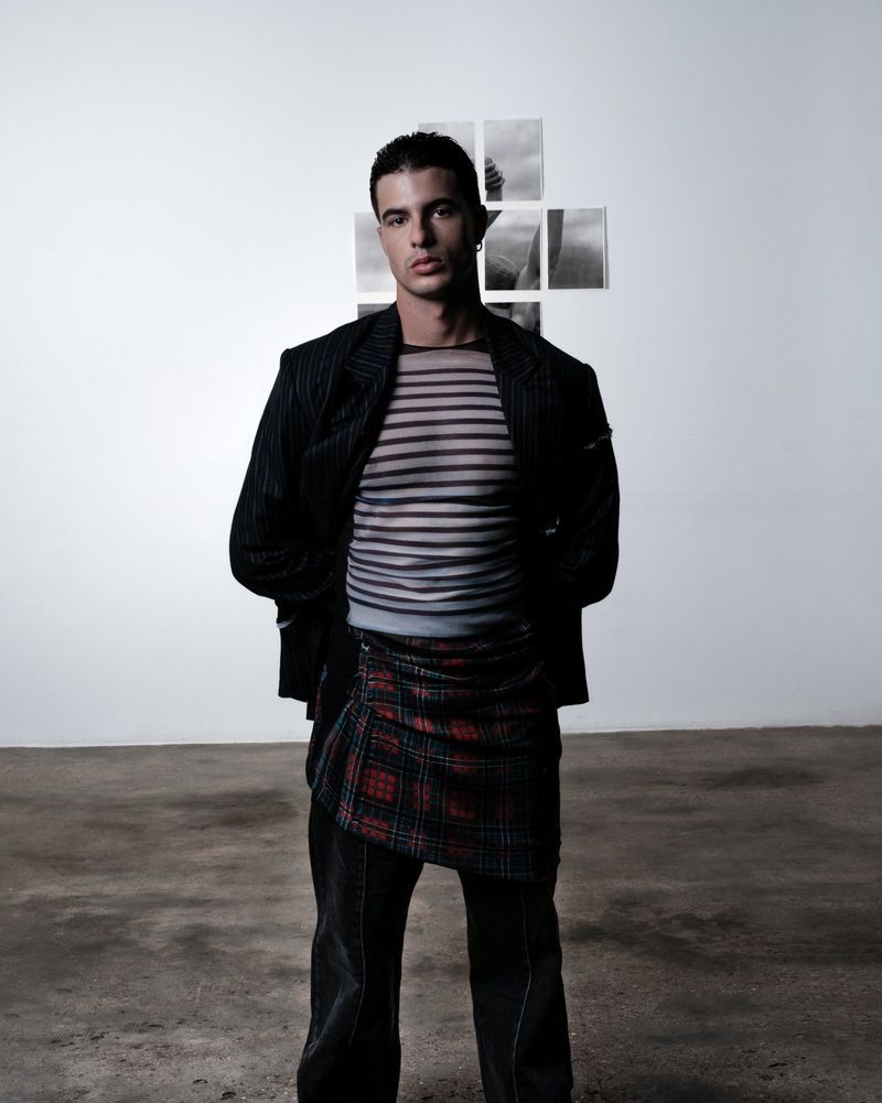 A young adult male stands in a photo studio wearing a sheer striped shirt, blazer, and patterned skirt with a white wall background.