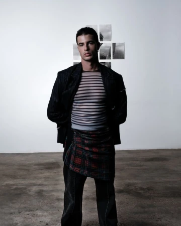 A young adult male stands in a photo studio wearing a sheer striped shirt, blazer, and patterned skirt with a white wall background.