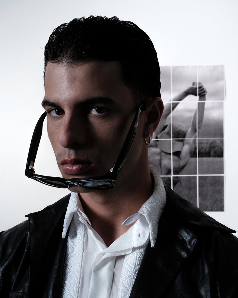Portrait of adult male wearing sunglasses with black jacket and white shirt in photo studio with framed art print background.