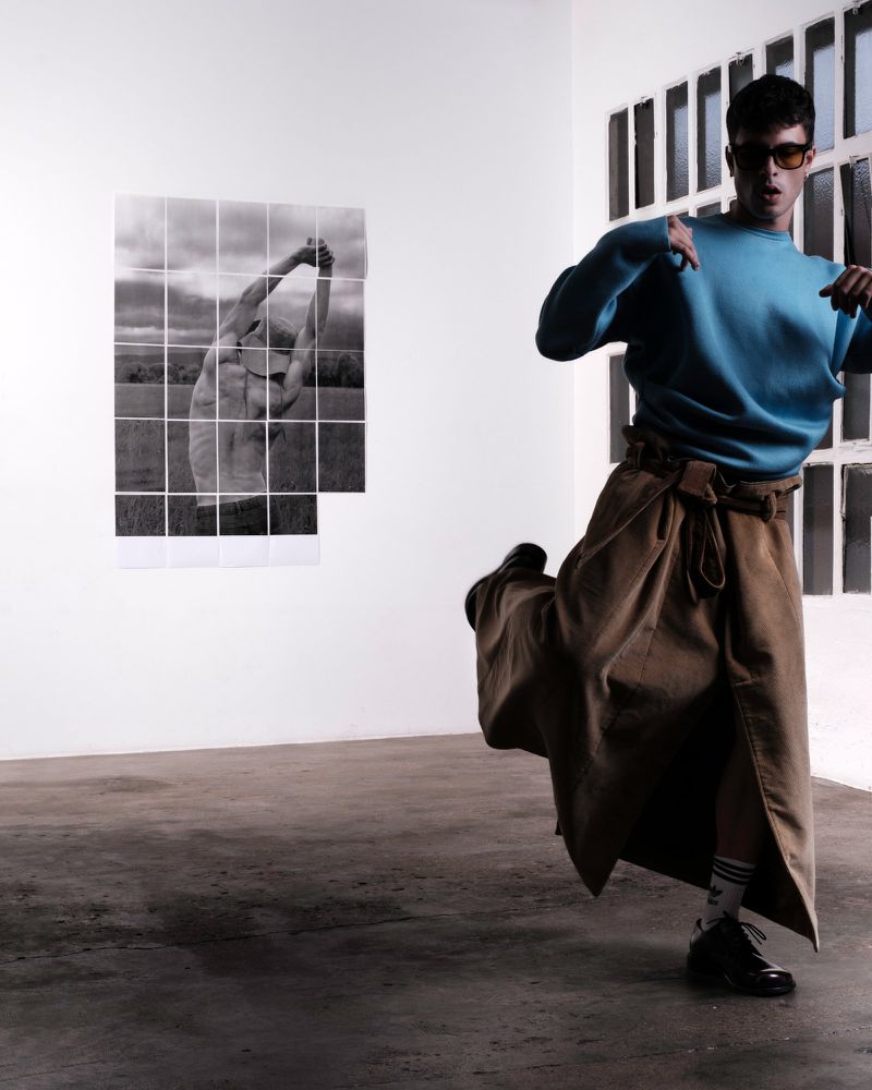 Male fashion model strikes a dynamic pose in a studio, wearing a blue sweater and tan skirt with an art print backdrop and indoor setting.