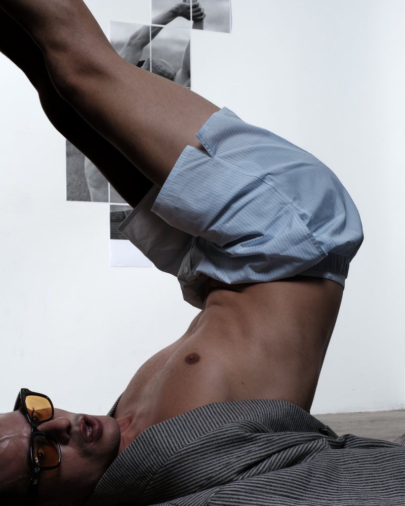 Male figure lying with legs up, blue striped shorts, gray shirt, sunglasses; monochrome images in background; studio lighting.