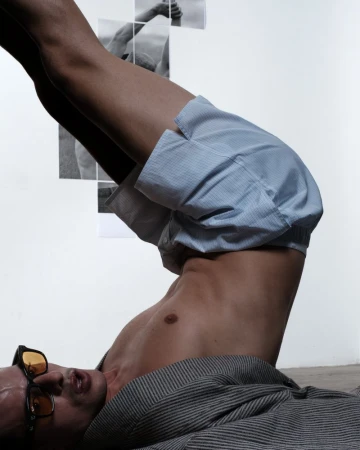 Male figure lying with legs up, blue striped shorts, gray shirt, sunglasses; monochrome images in background; studio lighting.