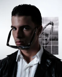Portrait of a man in a white shirt and black jacket with glasses held in his mouth against a white wall.