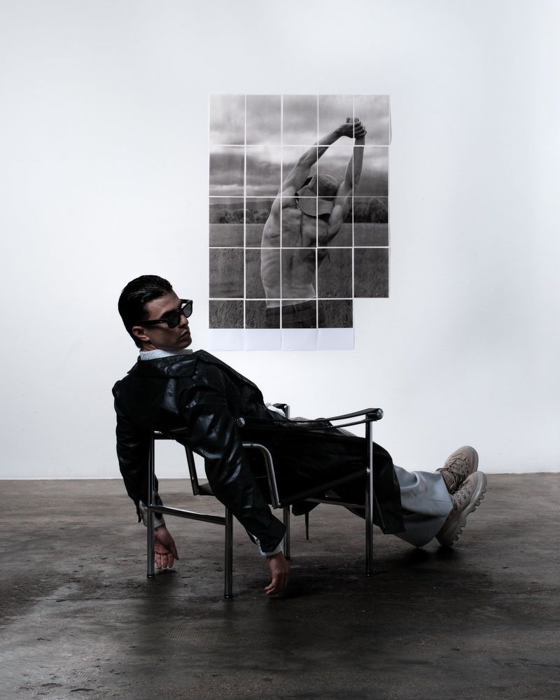 Man in a black leather jacket and sunglasses reclining in a chrome armchair in front of a tiled photo collage on a white wall.