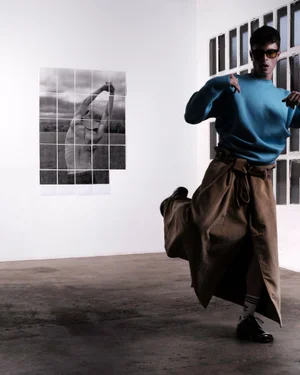 Man wearing blue sweater and tan skirt dancing in a white room with a photo collage on the wall