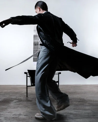 Man spinning in a long flowing black leather trench coat and grey wide-leg trousers in a minimalist studio setting.