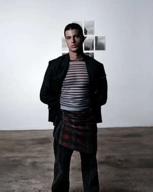 Portrait of a man wearing a striped sheer top and dark jacket against a plain wall.