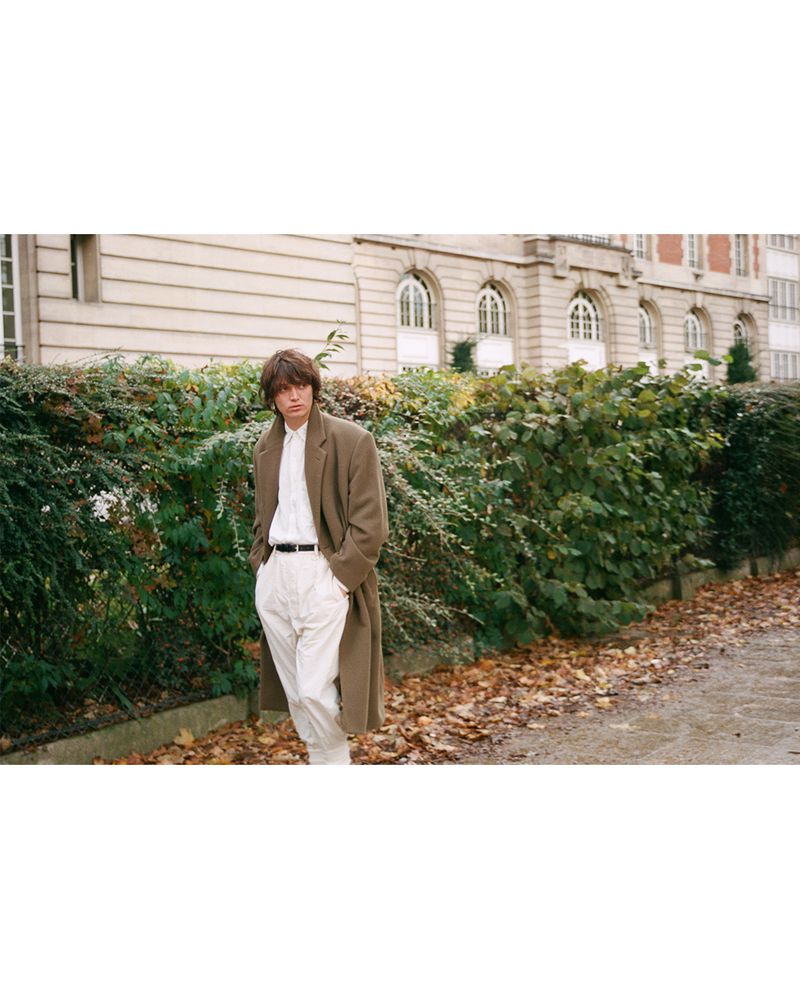 Man in long coat poses in front of a building and green foliage. He wears a white shirt and light-colored trousers, outdoors.