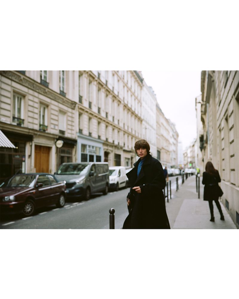 Young adult in a black coat stands on a sidewalk in an urban setting with beige buildings and parked cars in the background.