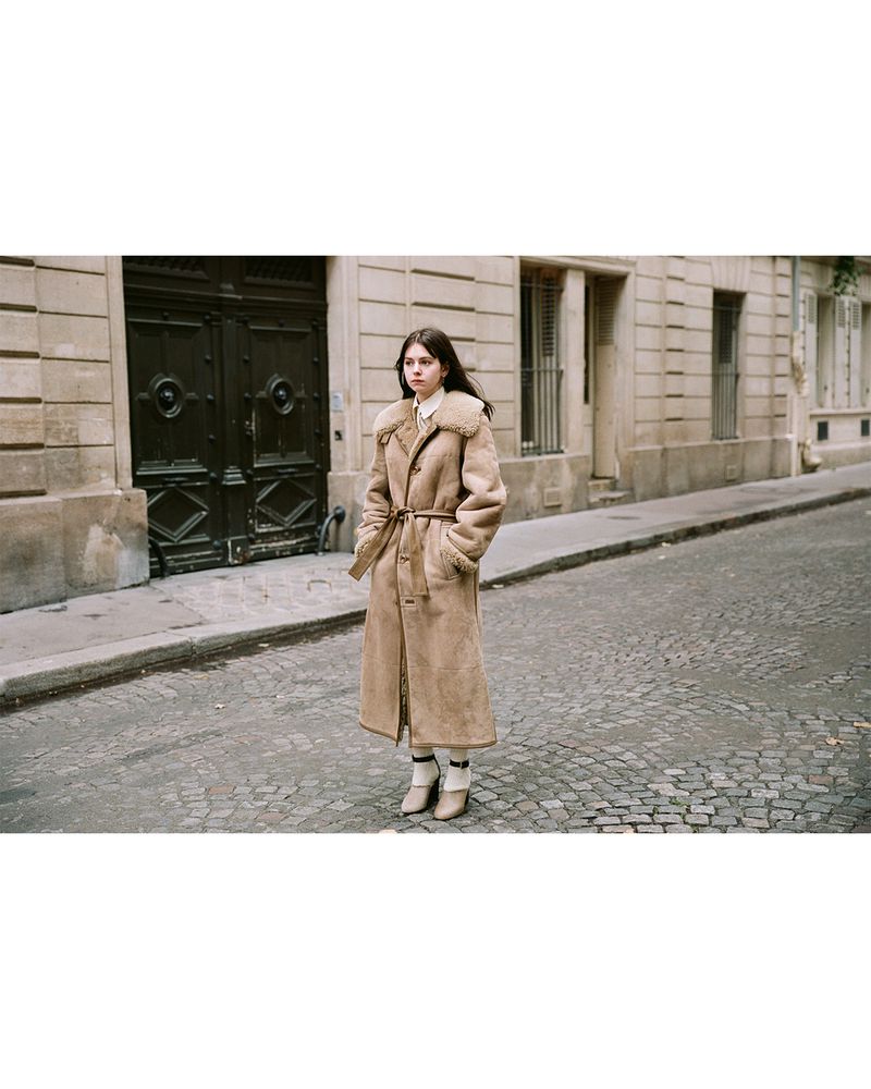 Young woman in a tan shearling belted coat posing in an urban setting with cobblestone street and stone buildings in the background.