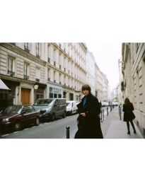 Model in a tailored long black overcoat and blue shirt looking over his shoulder while walking along a city sidewalk.