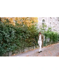 Model wearing a long brown coat and white trousers walking along an autumn street with lush foliage and a residential building facade.