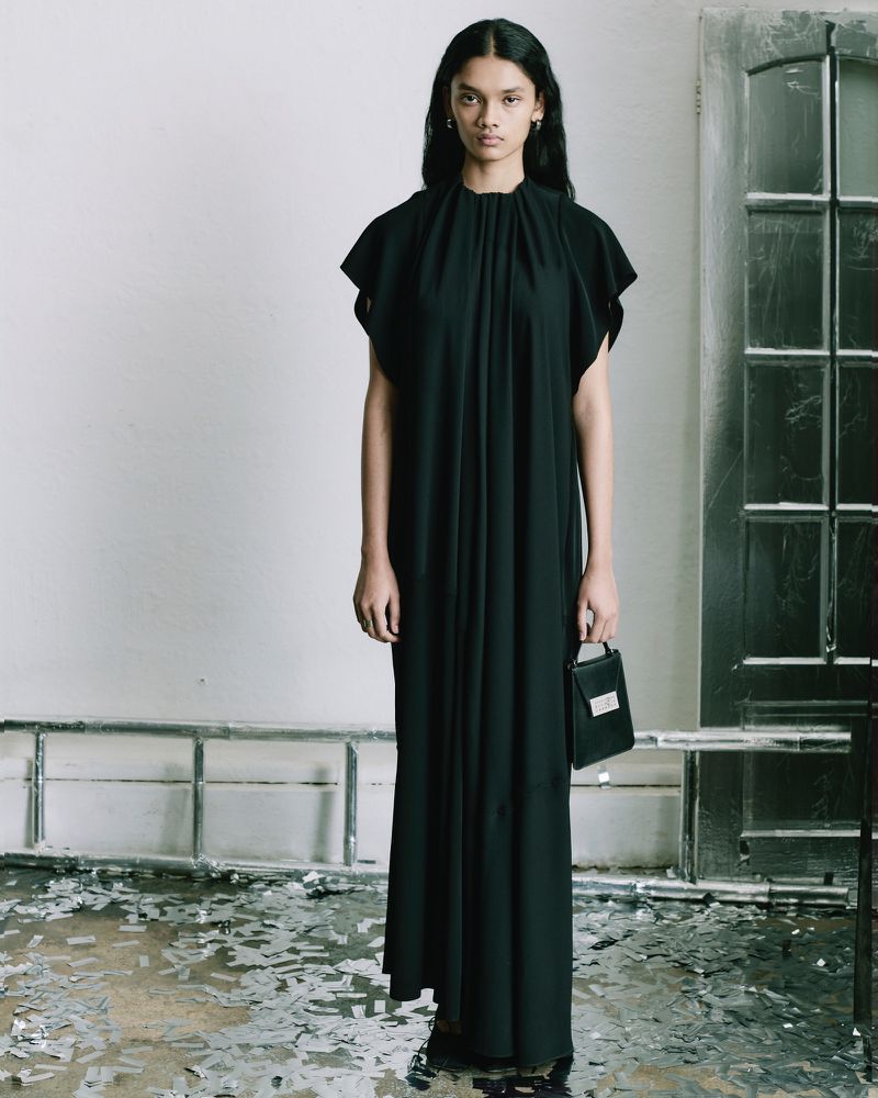 A solo, androgynous model showcases a floor-length black dress and a small handbag in a minimally adorned, neutral studio environment.