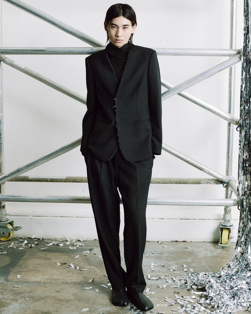 A person with long dark hair poses in an all-black suit against a backdrop of scaffolding and scattered silver material.