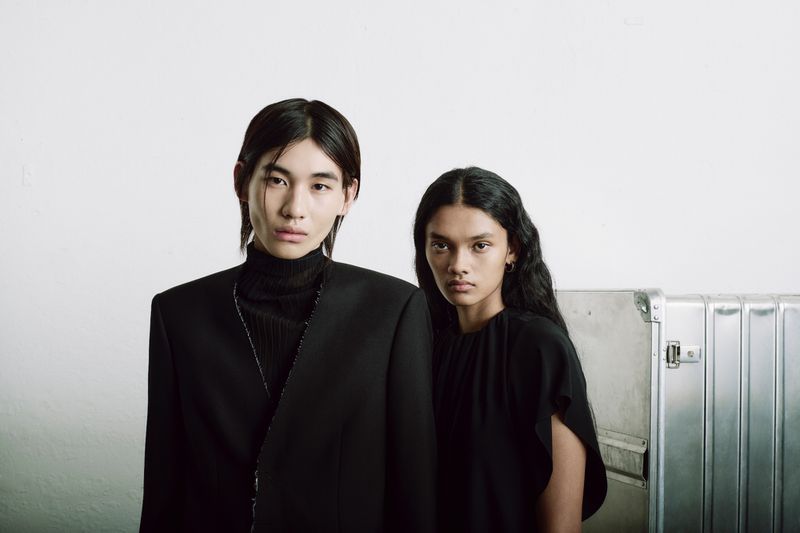 Two fashion models in black clothing pose in front of a textured white wall with a metallic case. Moody lighting and a studio setting.